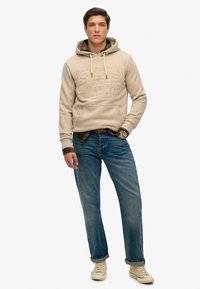 Beige hoodie met reliëflogo, geblokte voering, denim jeans, opgerolde mouwen en beige sneakers. Casual, relaxed fit, katoenmix materiaal.