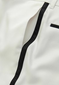 Tissu blanc avec un motif texturé, featuring un passepoil noir le long de la couture et une petite poche noire en accent.