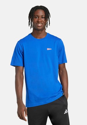 T-shirt en coton bleu à manches courtes et encolure ronde. Présente un petit logo en forme de triangle rouge sur la poitrine. Associé à un pantalon noir.
