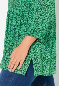 Blusa verde con motivo leopardato, maniche corte, corps smockato, fessure laterali e tessuto leggero. La indossatrice tocca l'orlo.