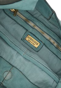 Interno di una borsa a tracolla in pelle teal con una patch del logo in oro, due scomparti con zip e una texture liscia con sottili dettagli di cucitura.