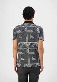 Polo de manga corta con un patrón geométrico en blanco y negro, que presenta líneas diagonales y formas variables, combinado con jeans oscuros.