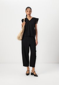 Top nero a V con maniche arricciate, abbinato a pantaloni neri larghi. Borsa beige intrecciata e scarpe nere con zeppa e cinturino.