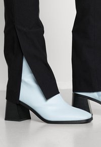 Botas de tornozelo de couro azul claro com bicos pontudos e saltos grossos com listras pretas, combinadas com calças de tecido pretas com aberturas.