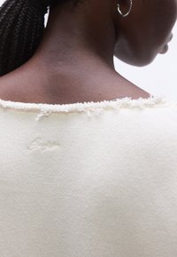 Gros plan sur le cou et le haut du dos d’une personne portant un haut couleur crème avec un col effiloché et un texte brodé sur le tissu.