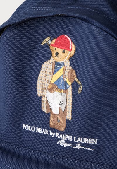 Κεντημένη αρκουδάκι που φοράει κόκκινο κράνος, καρό παλτό, μπλε πουκάμισο, λευκό παντελόνι, κρατώντας ρακέτα πόλο σε φόντο από σκούρο μπλε ύφασμα.