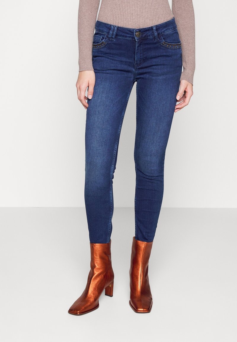 Femme portant un jean skinny bleu foncé avec des détails de poches cloutées et des bottines métalliques cuivre, debout devant un fond uni.