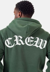 Tmavo zelená mikina s kapucňou vyrobená z mäkkej látky, na ktorej chrbte je biele písmo v gotickom štýle s nápisom "CREW". Nastaviteľná kapucňa s šnúrkou.