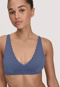 Jonge vrouw die een blauwe zachte beha draagt met een scalloped kanten rand en brede schouderbanden, zichtbaar van de borst omhoog tegen een effen achtergrond.
