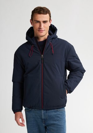 Chaqueta acolchada de color azul marino con capucha, cordones rojos, cremallera frontal y puños elásticos, combinada con jeans de color azul claro.