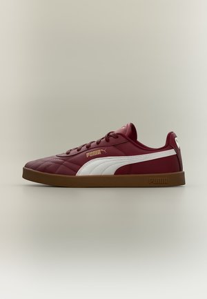 Μαρόν αθλητικό παπούτσι Puma με λευκή ρίγα, χρυσό λογότυπο Puma, σχεδιασμό με κορδόνια και καφέ λαστιχένια σόλα σε απλό ανοιχτό φόντο.