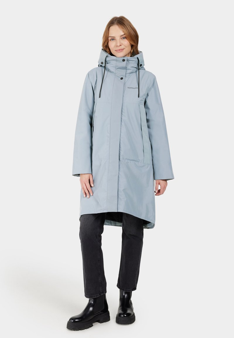 Helle blaue wasserdichte Jacke mit hohem Kragen, Frontreißverschluss, verstellbaren Kordelzügen und Seitentaschen, kombiniert mit schwarzen Slim-Fit-Jeans und Stiefeln.