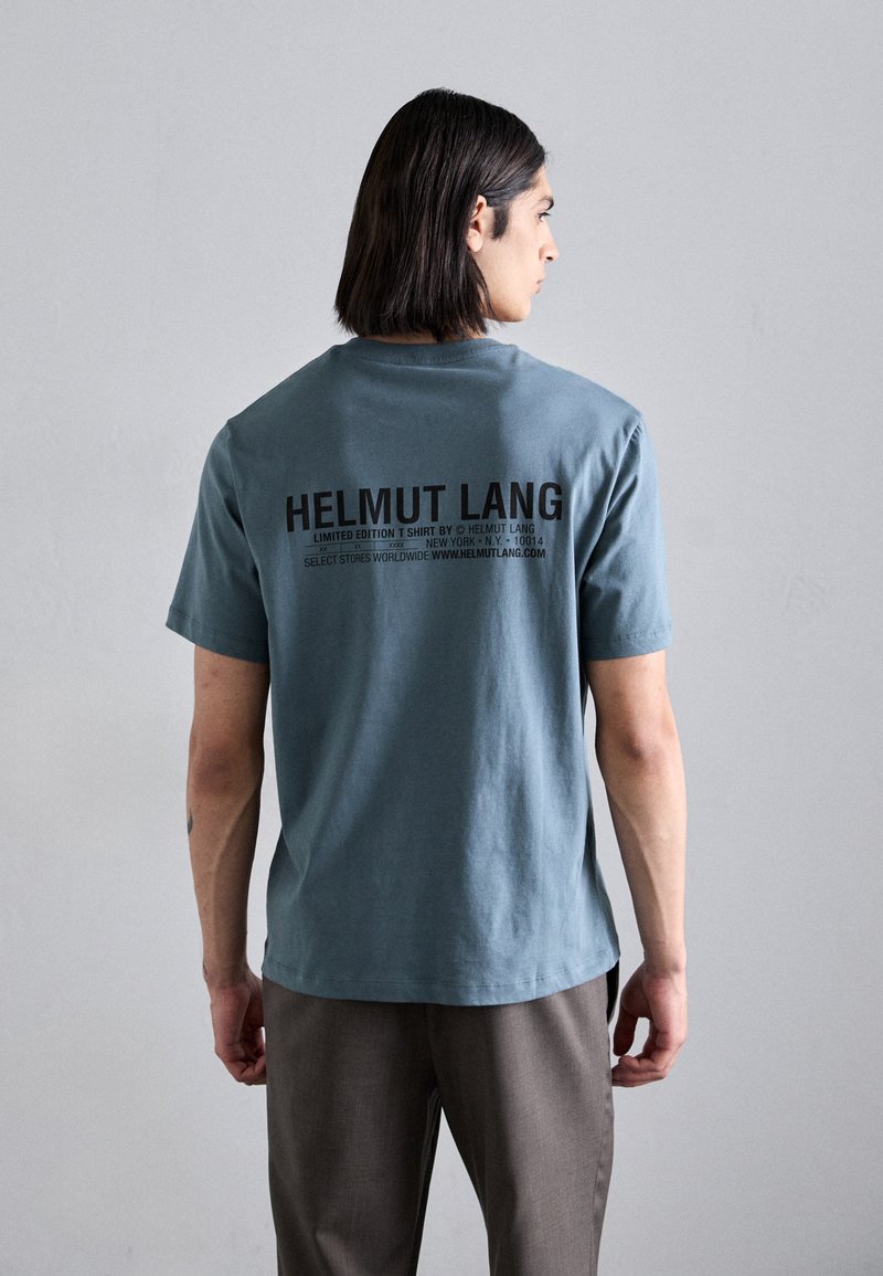 Himmelblå t-shirt med rund halsringning, korta ärmar och en svart tryckt logotyp på baksidan. Materialet verkar mjukt och avslappnat.
