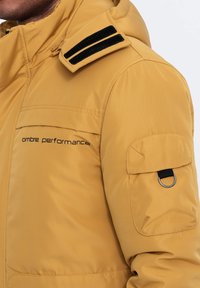 Gul vadderad jacka med dragkedja, axelremmdetalj och en sidoficka med en dekorativ ring, märkt med "ombre performance."