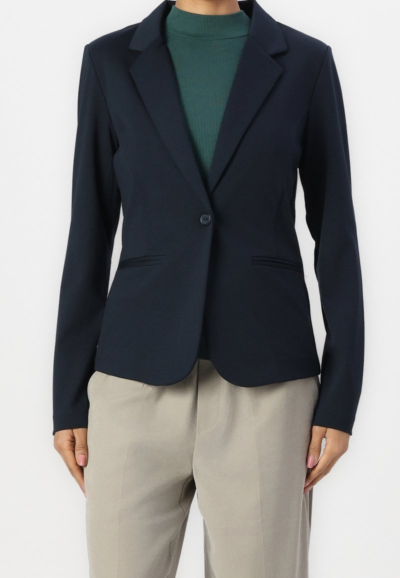 Veste de costume bleu marine à coupe ajustée, fermeture à un bouton, revers crantés et deux poches avant, portée sur un haut côtelé vert et un pantalon beige.