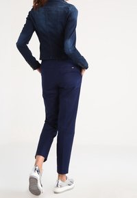 Chaqueta de mezclilla en azul oscuro con un diseño ajustado, combinada con pantalones azul marino y sneakers plateados. Enfoque en la estructura y el ajuste de la ropa.