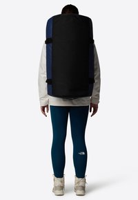 Zaino nero e blu con spalline regolabili, forma rettangolare, esterno in tessuto. Persona che indossa una giacca nera e leggings turchesi, stivali bianchi.