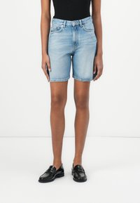 Shorts de mezclilla en azul claro, con un corte clásico, cinco bolsillos, trabillas para cinturón y un dobladillo ligeramente desgastado. Llevados con zapatos de charol negros.