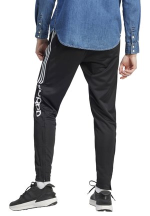 Pantalones deportivos negros con rayas y logo blancos, fabricados en tejido suave. Presentan bolsillos con cremallera y un diseño ajustado en los tobillos.