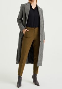 Dlouhý šedý kabát s houndstooth vzorem přes černou košili, kombinovaný s olivově zelenými kalhotami a šedými koženými kotníkovými botami na podpatku. Kalhoty mají zapínání na knoflík.