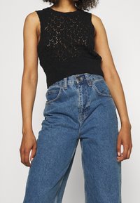 Schwarzes gestricktes ärmelloses Top mit einem strukturierten Muster, kombiniert mit hoch taillierten blauen Jeans im klassischen Fünf-Taschen-Design.