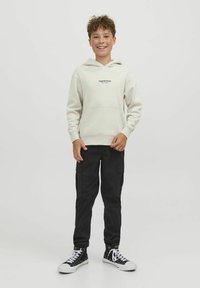 Felpa beige chiaro con tasca frontale e logo, abbinata a pantaloni cargo neri e sneakers alta nere, su uno sfondo neutro.