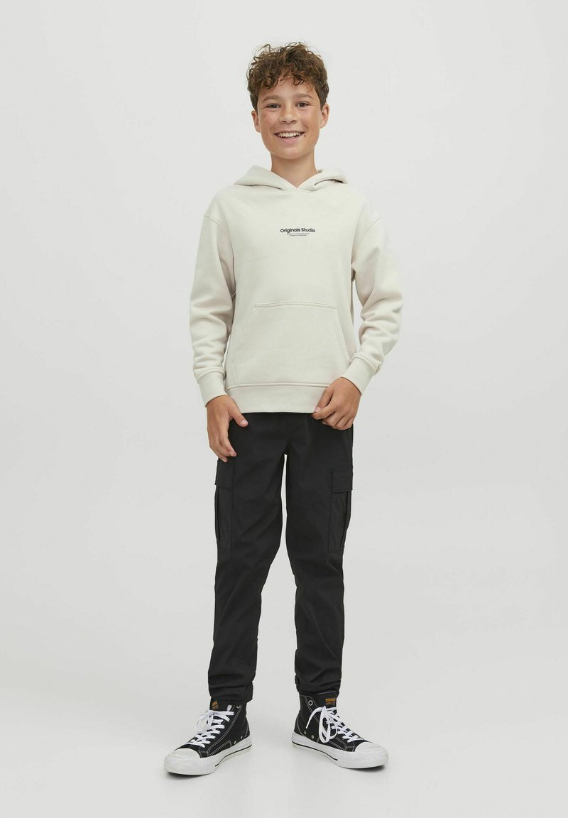 Felpa beige chiaro con tasca frontale e logo, abbinata a pantaloni cargo neri e sneakers alta nere, su uno sfondo neutro.