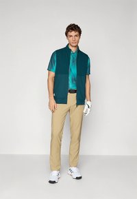 Golfoutfit med en tealärmlös väst över en blågrönmönstrad pikétröja, beige byxor och vita skor med svarta detaljer.