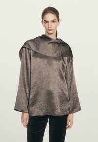 Metaliczna brązowa bluzka z szerokim, opadającym kołnierzem i długimi rękawami. Teksturowana tkanina ma lekko pomarszczony wygląd.
