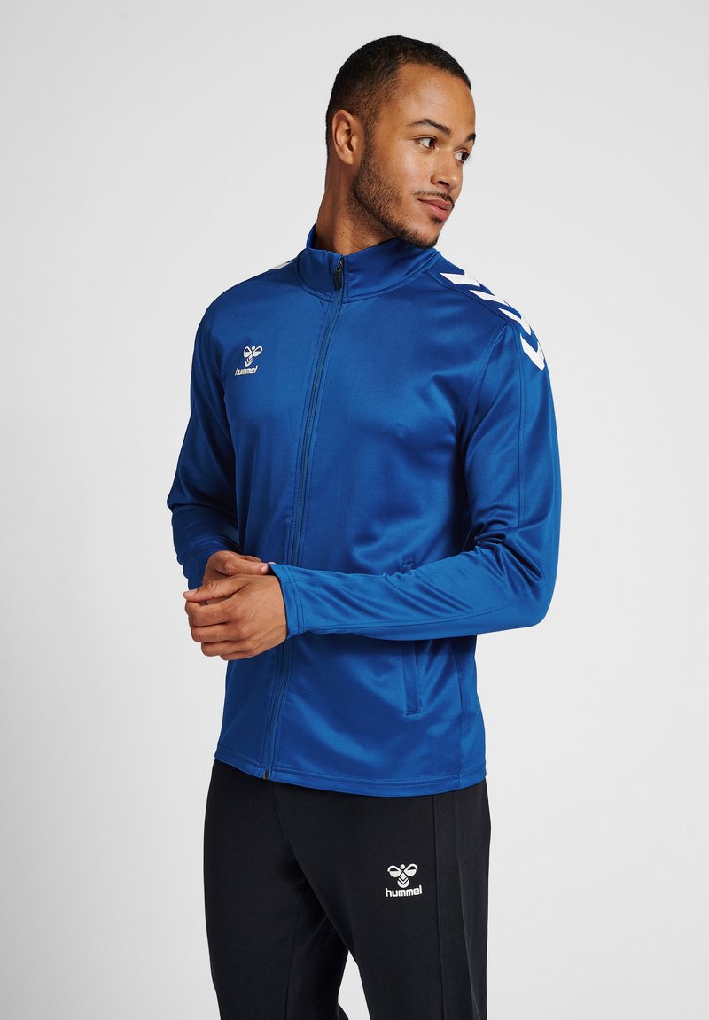 Blue athletic jacket with long sleeves, zip front, and white shoulder accents. Made of smooth fabric, paired with black pants featuring logo.