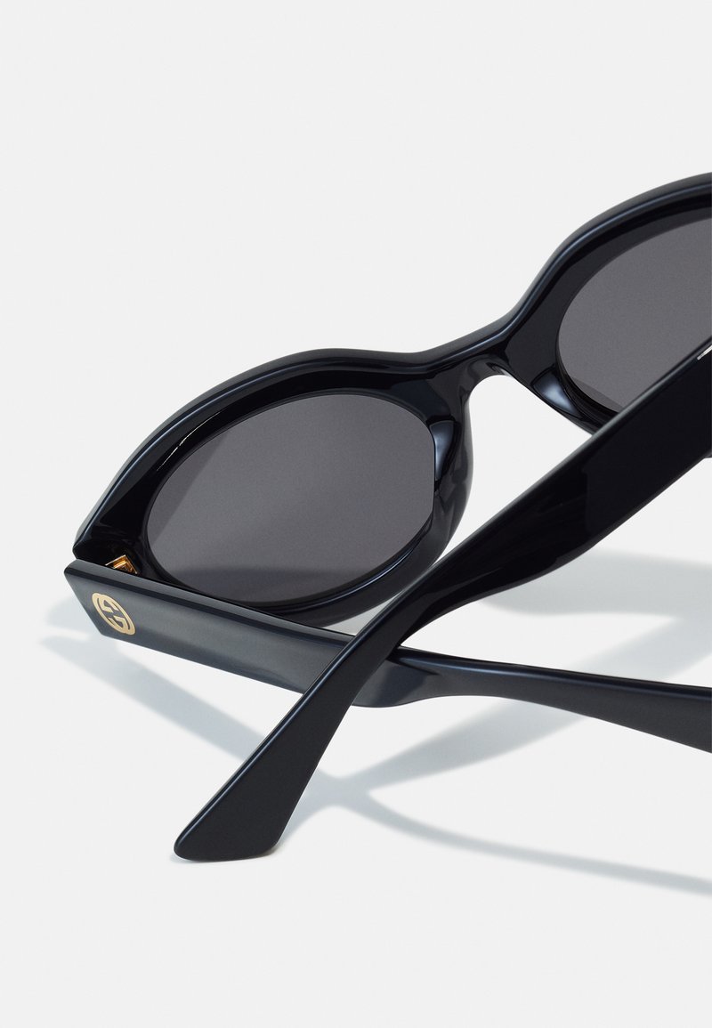 Black sunglasses with a sleek, glossy finish. Features large oval lenses and narrow, elongated temples. Gold logo accent on the side.