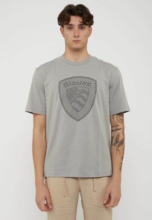 Giovane uomo con capelli corti e ricci che indossa una maglietta grigia con un grande stemma a scudo decorato e pantaloni beige, in piedi su sfondo bianco.