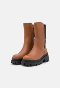 Une paire de bottes en cuir marron mi-mollet avec des semelles en caoutchouc noir épais et des zips sur les côtés, vue de face en angle sur fond blanc.