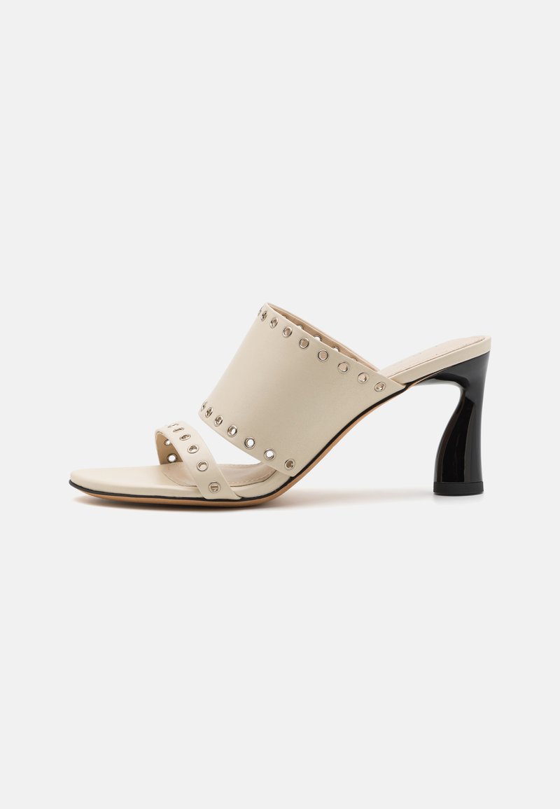 Beige hoge hakken mules met een brede band met zilveren studs, een smalle band over de voet en een glanzende zwarte taps toelopende hak.
