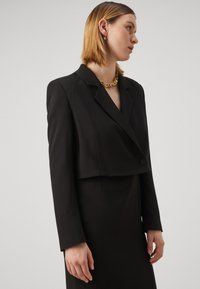 Crna skraćena blejzer jakna s izrezanim kragnom, kopčanjem na dugmad i dugim rukavima. Uparen s elegantnom crnom haljinom koja ima glatku teksturu.