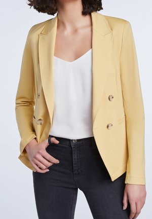 Vrouw draagt een lichtgele dubbelgeknoopte blazer, een wit topje met V-hals en donkere strakke jeans, met één hand in de zak.