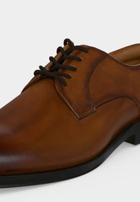 Chaussure habillée en cuir marron avec lacets noirs et semelle noire, vue en gros plan de côté sur fond blanc.