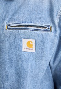 Lichtblauwe spijkerjas met een ritszak op de borst. Kenmerkt zich door een vierkant Carhartt-label met een geel logo en rode tekst.