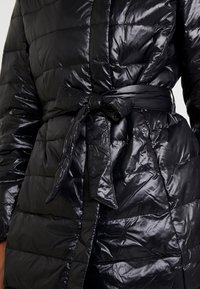 Abrigo acolchado negro con un acabado brillante, que cuenta con un cinturón de cintura atado. Secciones texturizadas y acolchadas crean un diseño estructurado.