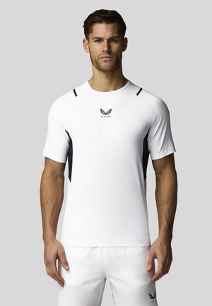 Homme portant un t-shirt de sport blanc et un short avec des accents noirs et un petit logo ailé sur la poitrine et la jambe, se tient devant un fond uni.