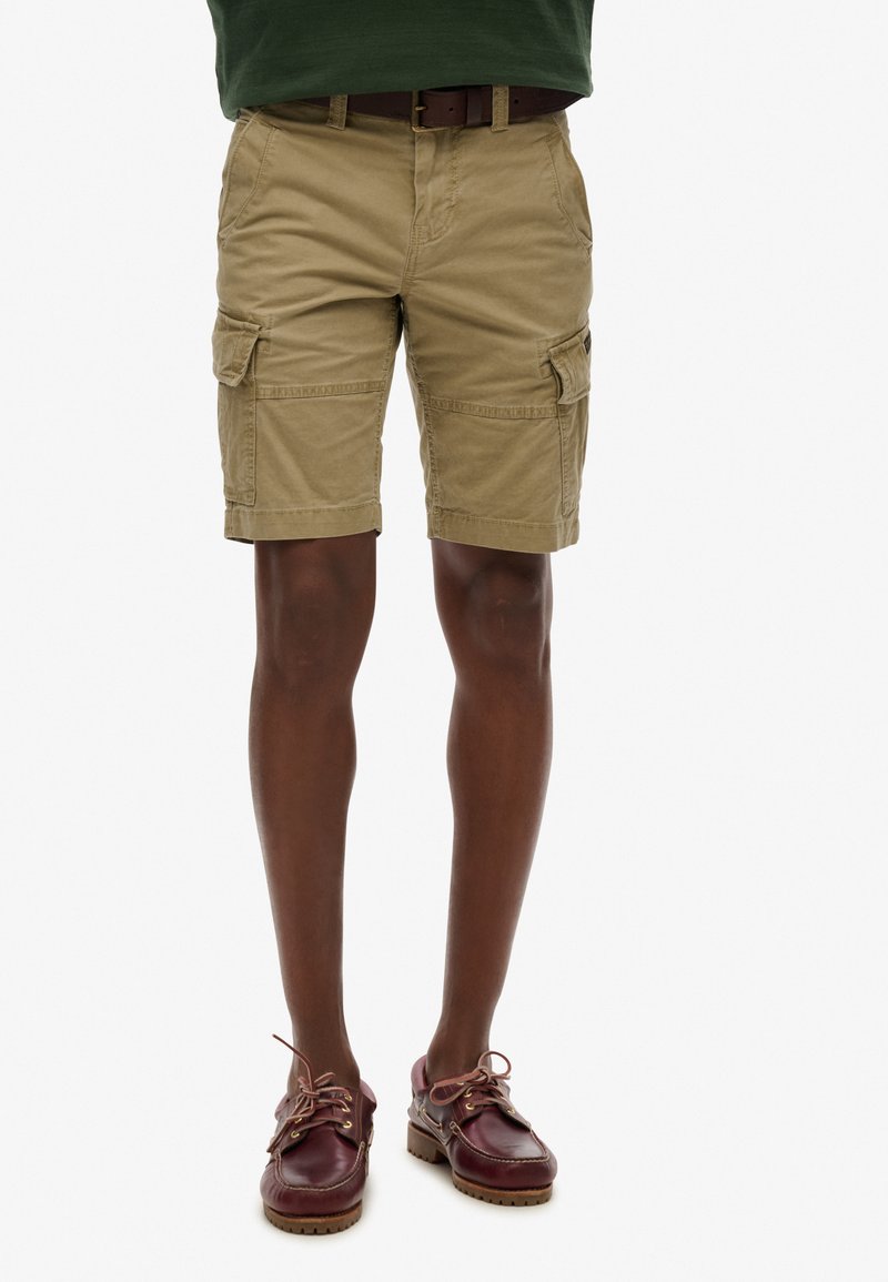 Pantaloni cargo color khaki, dotati di tasche laterali e dallo stile rilassato. Indossati con scarpe marroni, abbinati a una semplice cintura in pelle.