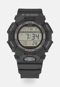 Cyfrowy zegarek G-Shock z czarnym paskiem z żywicy, kwadratową tarczą, dużymi przyciskami i wyświetlaczem LCD pokazującym godzinę, datę oraz różne funkcje. Wodoodporny do 20 bar.