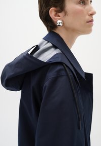 Jūras zila kapucejuma jaka ar strukturētu apkakli, gludu iekšējo oderi un izteiksmīgu melnu auklas detaļu uz pleca.