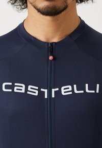 Le maillot de cyclisme bleu marine présente une fermeture éclair intégrale, un logo "castelli" blanc bien en vue, et un tissu léger et texturé. Détail de tirette rouge.