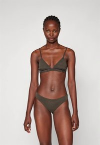 Femme se tenant devant un fond simple, portant un bikini vert olive foncé avec des fines bretelles et un haut en forme de triangle.