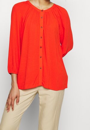Blusa leggera rossa a maniche lunghe con scollatura rotonda, chiusura con bottoni e dettagli arricciati sulle spalle. Accoppiata con pantaloni beige.