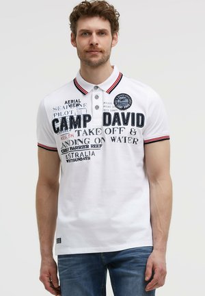 Man wearing white polo shirt with red and black striped collar and sleeves, printed text and patch, paired with blue jeans.