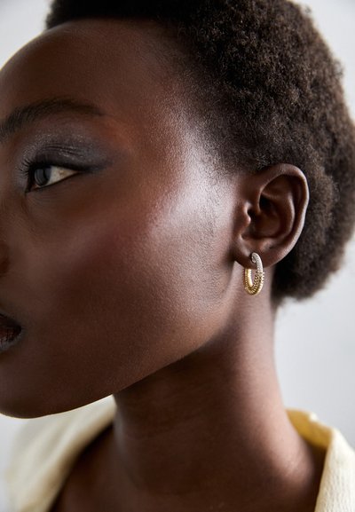 Gold- und Silbercreolen mit texturierten Oberflächen; das Modell hat kurzes, fest eingedrehtes Haar und eine glatte Haut; dezent geschminkt, hebt das Makeup die Gesichtszüge hervor.