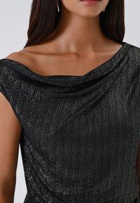 Donna che indossa un abito aderente nero con spalline scese e scollo drappeggiato, ripresa dalle spalle fino a metà torace.
