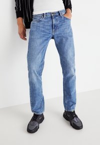 Vaqueros de mezclilla azul claro en un diseño de corte recto, que cuentan con un estilo clásico de cinco bolsillos y una sutil textura desgastada. Combinados con zapatillas negras.